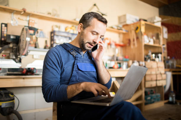 Portrait of mature worker using laptop and speaking by smartphone in workshop interior, copy space